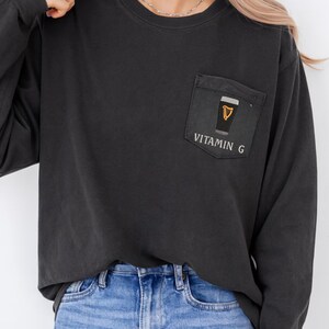 May include: Black long-sleeve shirt with a pocket featuring a graphic of a pint of beer with a gold harp and the text "VITAMIN G". The shirt is paired with blue jeans.