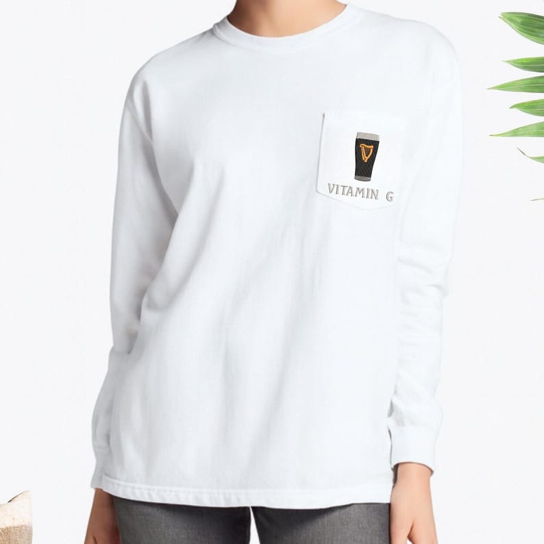 May include: White long-sleeve shirt with a pocket featuring a graphic of a pint of stout beer. The beer has a black body and a tan head, with the text "VITAMIN G" below the pint.