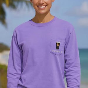 May include: A long-sleeved, lavender shirt with a pocket featuring a graphic of a pint of stout with a gold harp. The text "Vitamin G" is below the graphic. The shirt is a solid color and has a crew neck.