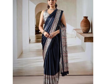May include: A dark navy blue satin saree featuring a wide, intricate border with a repeating pattern in shades of cream, red, and black. The saree is draped over a matching sleeveless blouse with a high neckline and a metallic silver choker necklace.