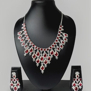 May include: A silver necklace and earring set with red, teardrop-shaped gemstones. The necklace has a cascading design with multiple gemstones, and the earrings mirror the necklace's style. The jewellery is displayed on a black mannequin.