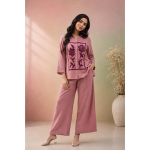 Peut inclure: Un ensemble deux pièces rose poudré composé d'une chemise à manches longues boutonnée et d'un pantalon large. La chemise présente un empiècement rectangulaire violet foncé sur le devant avec un motif brodé de deux roses sur tiges.