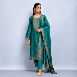 May include: A teal silk-blend kurta set featuring a long-sleeved tunic with a gold embroidered neckline and hem, matching straight-leg trousers, and a coordinating teal dupatta with a wide gold border, accented by gold drop earrings.