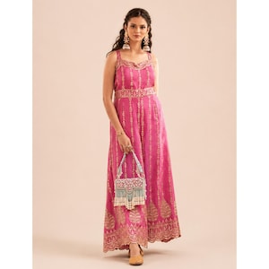 May include: A vibrant fuchsia jumpsuit featuring a sleeveless bodice with thin straps and a wide-leg silhouette. The garment is adorned with intricate gold-toned vertical patterns and leaf motifs. A small, rectangular, light blue beaded handbag is held.