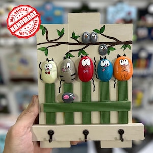 Puede incluir: Llavero de madera hecho a mano con diseños de gatos y pájaros pintados. El llavero tiene un fondo crema con una valla verde, una rama con pájaros y seis figuras coloridas con forma de gato. El llavero tiene tres ganchos en la parte inferior y el texto "100% Original Handmade 100% Original".