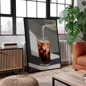 May include: A framed print depicts a tall glass of iced coffee with ice cubes and milk being poured in. The coffee is dark brown, and the milk creates a swirling pattern. The background is a neutral gray.