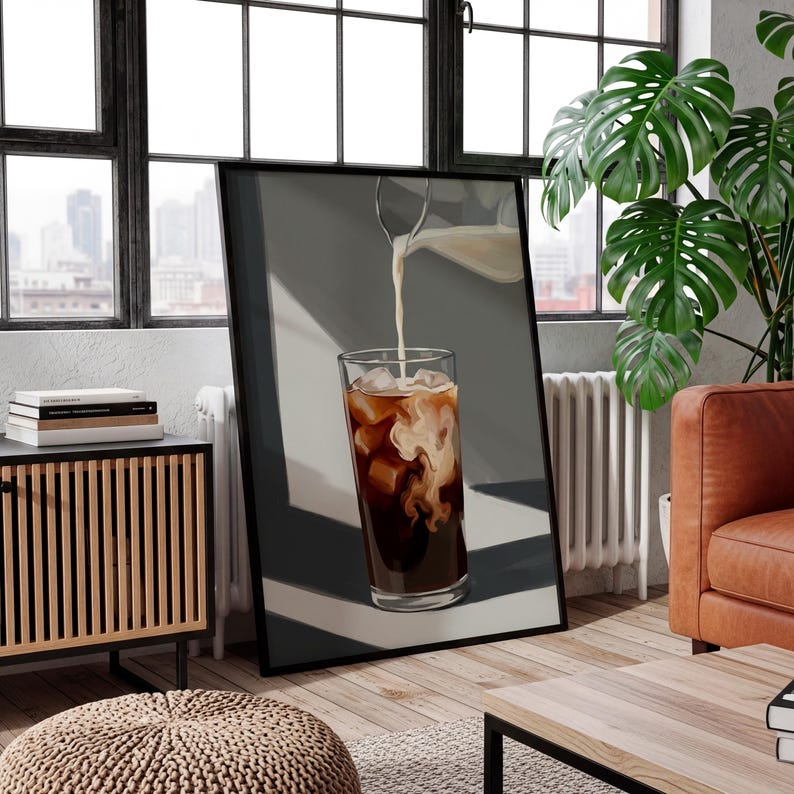May include: A framed print depicts a tall glass of iced coffee with ice cubes and milk being poured in. The coffee is dark brown, and the milk creates a swirling pattern. The background is a neutral gray.