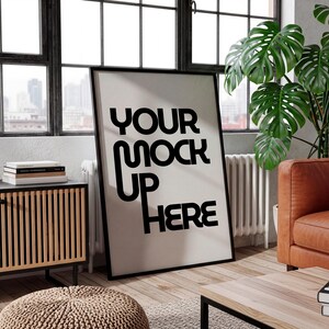 May include: A large black framed poster with the text "YOUR MOCK UP HERE" in bold black letters. The poster is leaning against a wall in a room with a window, a leather couch, and a wooden cabinet.