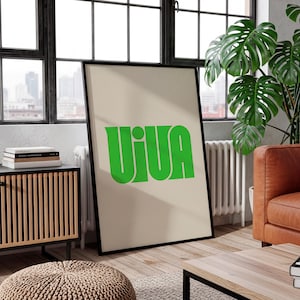 May include: A framed poster with the word "VIVA" in bold, green letters. The poster is set against a beige background and framed in black. The poster is leaning against a wall in a room with a window, a wooden cabinet, and a leather sofa.