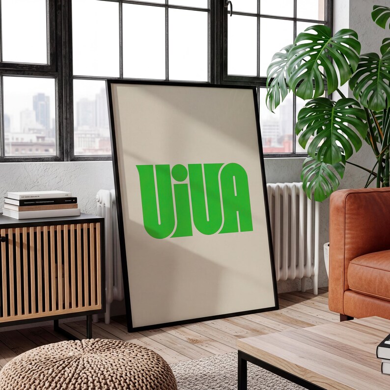 May include: A framed poster with the word "VIVA" in bold, green letters. The poster is set against a beige background and framed in black. The poster is leaning against a wall in a room with a window, a wooden cabinet, and a leather sofa.
