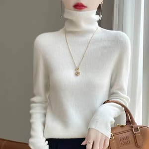 Op de afbeelding: Een witte coltrui met lange mouwen. De trui wordt gecombineerd met een gouden ketting met een hanger. Een bruine handtas met de merknaam "Miu Miu" is zichtbaar in de afbeelding.