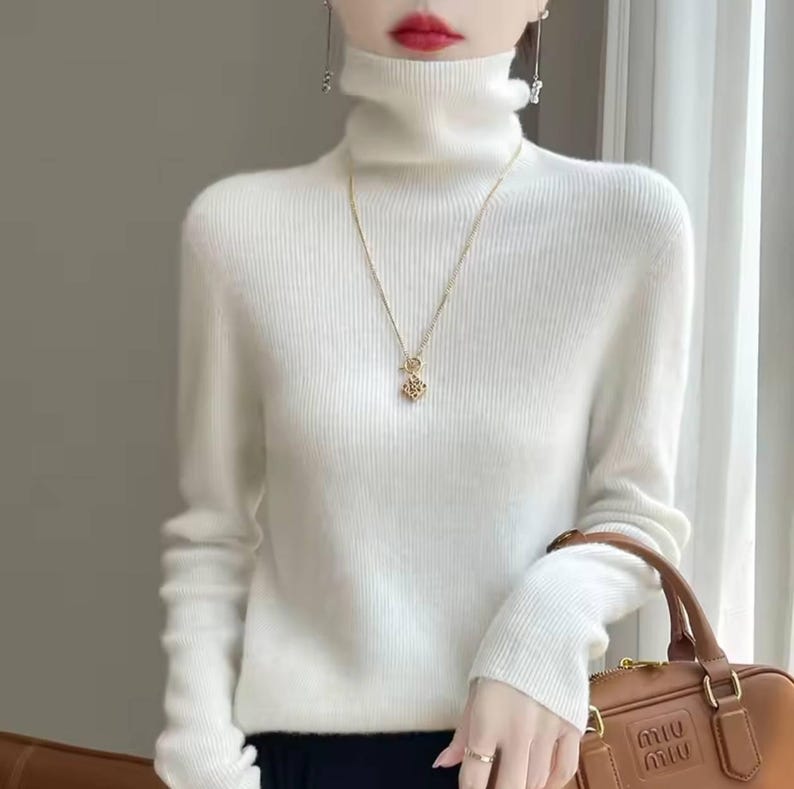 Op de afbeelding: Een witte coltrui met lange mouwen. De trui wordt gecombineerd met een gouden ketting met een hanger. Een bruine handtas met de merknaam "Miu Miu" is zichtbaar in de afbeelding.