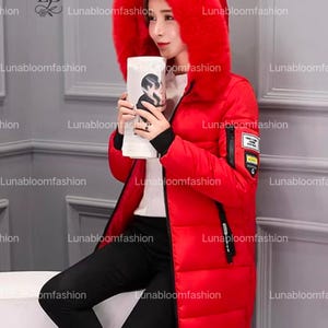 May include: A long, red winter coat with a large, fur-trimmed hood. The coat features a zipper closure, black accents, and a small patch on the sleeve. The model is holding a white object with a black and white image. The brand name "Lunabloomfashion" is visible.