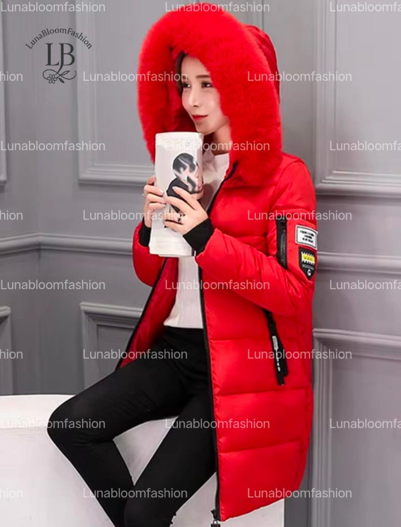 May include: A long, red winter coat with a large, fur-trimmed hood. The coat features a zipper closure, black accents, and a small patch on the sleeve. The model is holding a white object with a black and white image. The brand name "Lunabloomfashion" is visible.