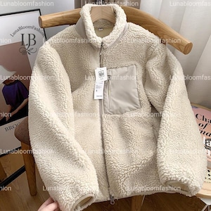 Puede incluir: Chaqueta de forro polar color crema con cuello alto y cremallera completa. La chaqueta presenta un bolsillo en el pecho de color beige claro y un exterior suave y texturizado. La chaqueta está colgada en una percha de madera.