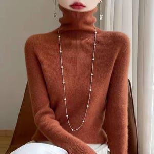 Op de afbeelding: Een roestkleurige coltrui met lange mouwen. Een delicate, lange ketting met kleine, ronde, witte parels hangt naar voren. De trui heeft een geribbelde textuur. Het model draagt bungelende oorbellen.