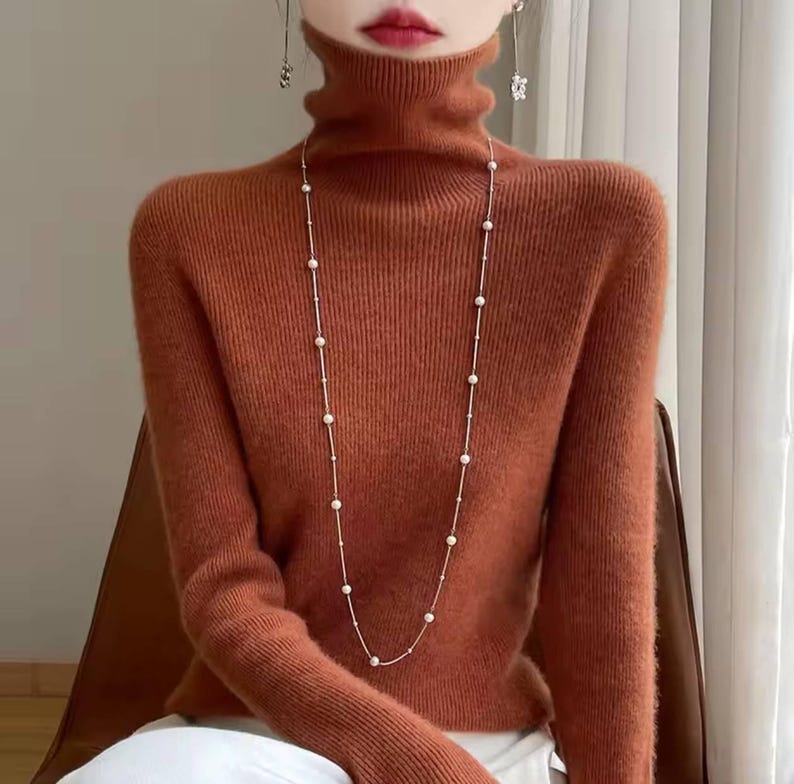 Op de afbeelding: Een roestkleurige coltrui met lange mouwen. Een delicate, lange ketting met kleine, ronde, witte parels hangt naar voren. De trui heeft een geribbelde textuur. Het model draagt bungelende oorbellen.