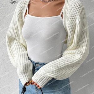 May include: A cream-coloured, chunky knit shrug sweater with long sleeves. The cropped bolero style is worn over a white vest top and blue jeans. The LunaBloomFashion logo is visible in the upper left corner.