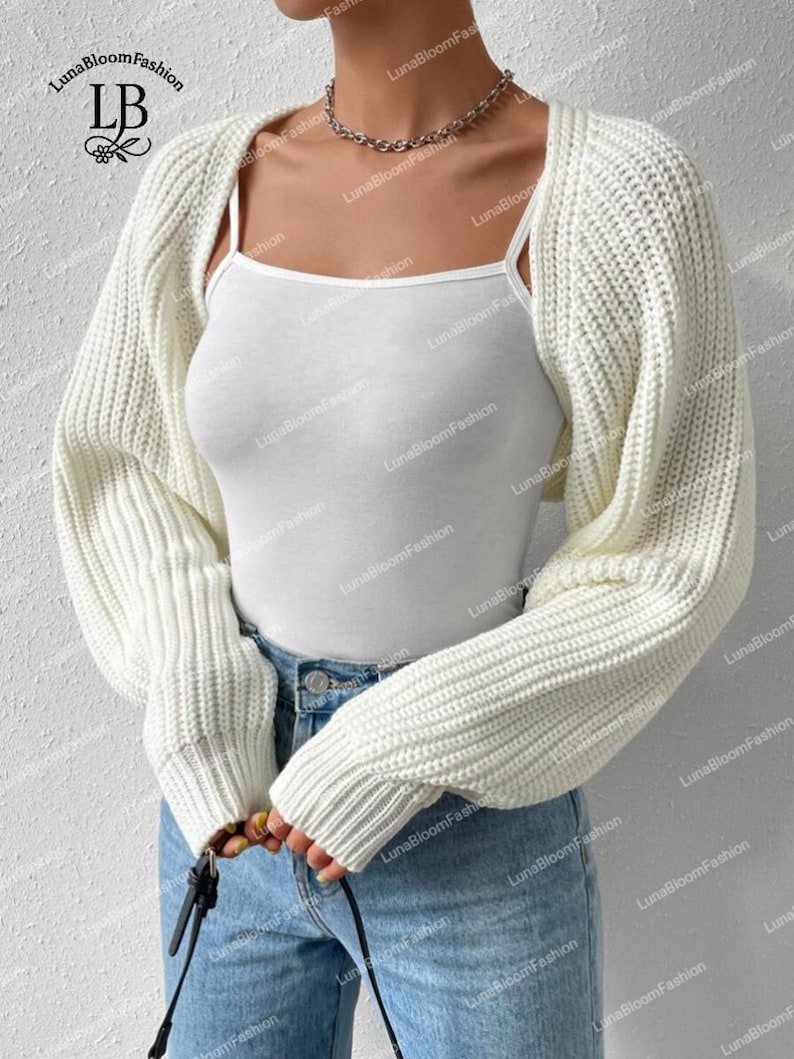 May include: A cream-coloured, chunky knit shrug sweater with long sleeves. The cropped bolero style is worn over a white vest top and blue jeans. The LunaBloomFashion logo is visible in the upper left corner.