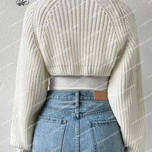 May include: A cream-coloured, cropped knit sweater with long sleeves. The sweater is worn with light blue denim jeans. The back view shows the ribbed texture of the sweater and the details of the jeans' pockets. The brand name "LunaBloomFashion" is visible.