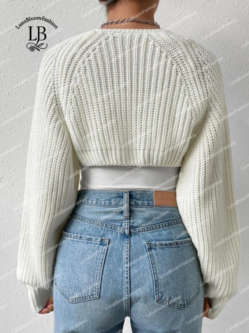 May include: A cream-coloured, cropped knit sweater with long sleeves. The sweater is worn with light blue denim jeans. The back view shows the ribbed texture of the sweater and the details of the jeans' pockets. The brand name "LunaBloomFashion" is visible.