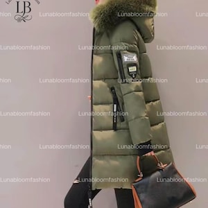 May include: Olive green puffer coat with a fur-trimmed hood. The long coat features a black zipper, a small patch on the sleeve, and a black handbag. The coat is designed for cold weather and is a stylish outerwear option.
