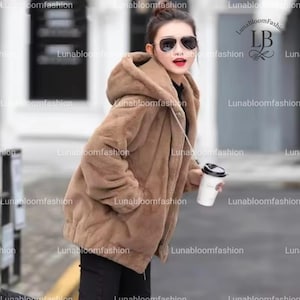 May include: A brown hooded faux fur coat with a zipper closure. The coat has a soft, plush texture and two front pockets. The model is wearing sunglasses and holding a coffee cup.