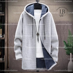 May include: A light gray, long-sleeved hooded jacket with a textured knit exterior and a dark blue, plush interior lining. The jacket features a full-length zipper, a drawstring hood, and a pocket on each side. A necklace with a pendant is visible.