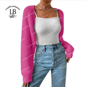 May include: A vibrant pink bolero-style sweater with long sleeves. The cropped sweater is made of a chunky knit fabric. The model is wearing a white vest top and blue jeans. The LunaBloomFashion logo is in the upper left corner.