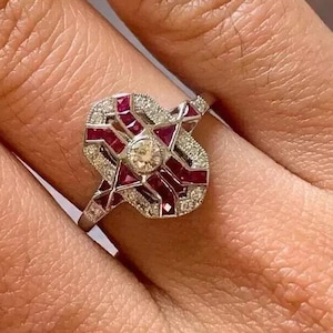 Può includere: Un anello d'argento con un design geometrico con pietre preziose rosse e piccoli diamanti. Una gemma centrale, rotonda e trasparente è circondata dal design. L'anello è su un dito.