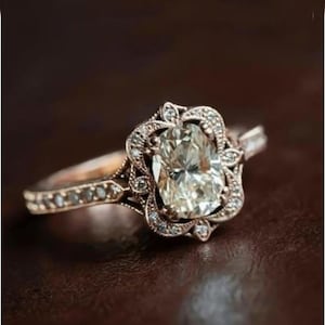 May include: A rose gold ring with a large, oval-cut, clear gemstone at the center. The gemstone is surrounded by smaller, clear stones and ornate filigree detailing. The band is also adorned with small stones.