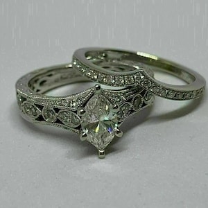Puede incluir: Un juego de anillos de compromiso de plata. El anillo de compromiso presenta un diamante central con talla marquesa y detalles intrincados. La banda que lo acompaña es curva y está engastada con pequeños diamantes.