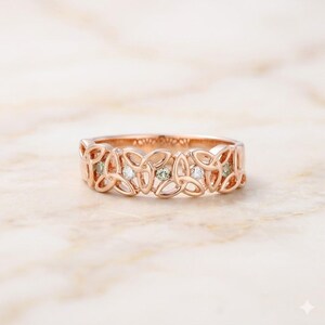 May include: A rose gold ring featuring an intricate openwork Celtic knot design that wraps around the entire band. The metal band is accented with small, round, clear and light green gemstones set within the repeating knot pattern.