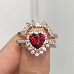 Puede incluir: Un conjunto de tres anillos con una piedra preciosa roja en forma de corazón. El conjunto incluye una banda con pequeñas piedras preciosas transparentes y un anillo en forma de corona con piedras de talla baguette. La piedra principal está rodeada por un halo de pequeñas piedras preciosas transparentes.