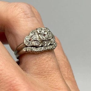 Op de afbeelding: Een vintage zilverkleurige verlovingsring en trouwring set. De verlovingsring heeft een vierkante centrale steen, omringd door kleinere stenen. De trouwring heeft een rij kleine stenen. De ringen worden aan een vinger gedragen.