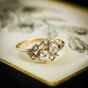 Puede incluir: Un anillo de estilo vintage con una banda de oro y un grupo de diamantes. El anillo presenta dos diamantes redondos más grandes como punto focal, rodeados de diamantes de acento más pequeños. El diseño tiene un detalle ornamental, tipo filigrana.