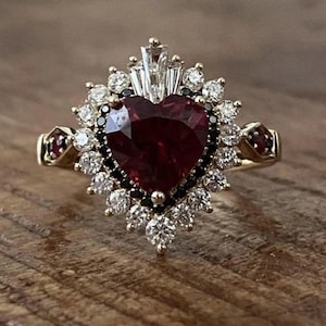 Könnte beinhalten: Ein goldener Ring mit einem herzförmigen, tiefroten Edelstein in der Mitte, umgeben von einem Halo aus weißen und schwarzen Edelsteinen. Das Design des Rings umfasst zusätzliche rote Edelsteine an den Schultern des Bandes.