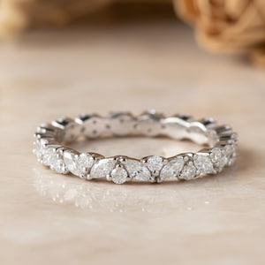 Könnte beinhalten: Ein silberner Eternity-Ring mit marquiseförmigen Diamanten, die um das Band gesetzt sind. Der Ring wird auf einer hellen Oberfläche mit verschwommenem Hintergrund präsentiert. Der Ring ist ein Schmuckstück.