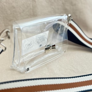 May include: A clear rectangular crossbody bag with a silver-toned metal turn-lock closure. The bag features a detachable woven shoulder strap with white, brown, and navy blue stripes. A small white rectangular label is visible inside the bag.