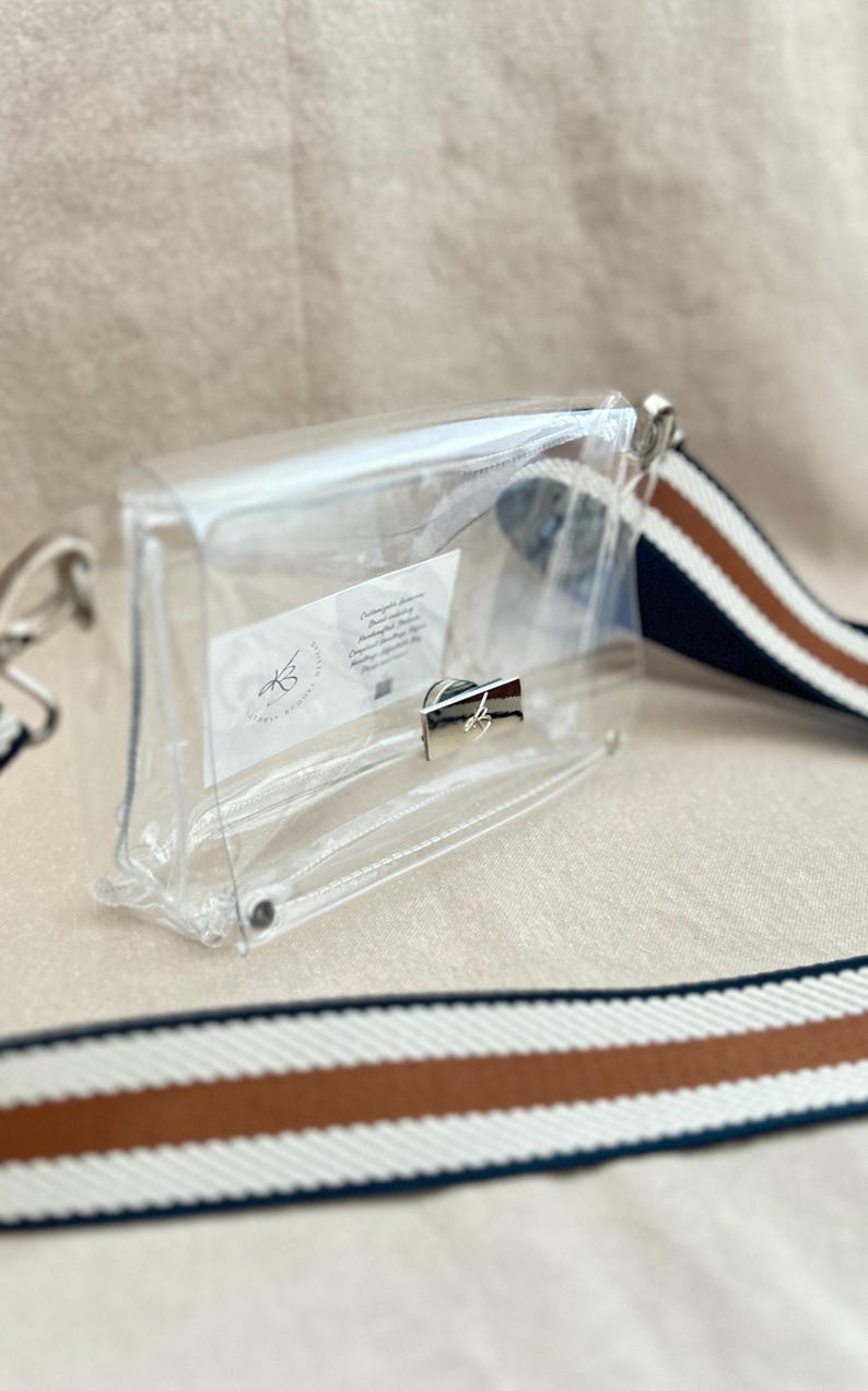 May include: A clear rectangular crossbody bag with a silver-toned metal turn-lock closure. The bag features a detachable woven shoulder strap with white, brown, and navy blue stripes. A small white rectangular label is visible inside the bag.