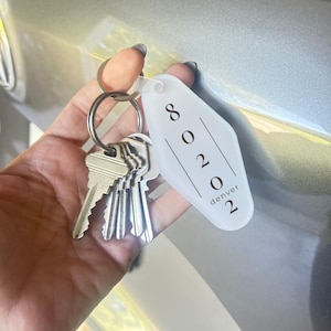 May include: A translucent white motel-style keychain with a diamond shape featuring the black printed text 80202 and denver. The keychain is attached to a silver metal split ring holding a cluster of four standard silver-colored metal keys.