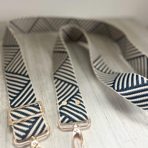 May include: A replacement bag strap featuring a woven geometric pattern in navy blue and cream. The strap includes adjustable gold-toned metal hardware, a sliding buckle, and two swivel lobster claw clasps attached to the ends with gold rivets.