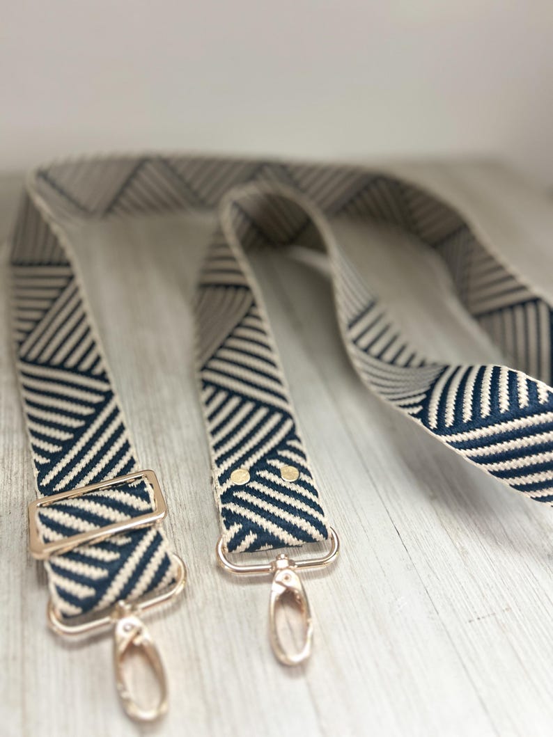 May include: A replacement bag strap featuring a woven geometric pattern in navy blue and cream. The strap includes adjustable gold-toned metal hardware, a sliding buckle, and two swivel lobster claw clasps attached to the ends with gold rivets.