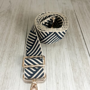 May include: A woven bag strap featuring a navy blue and cream chevron pattern. The strap includes a gold-toned metal adjustable slider and a swivel lobster clasp hook at the end, with the remaining length coiled into a circular shape.