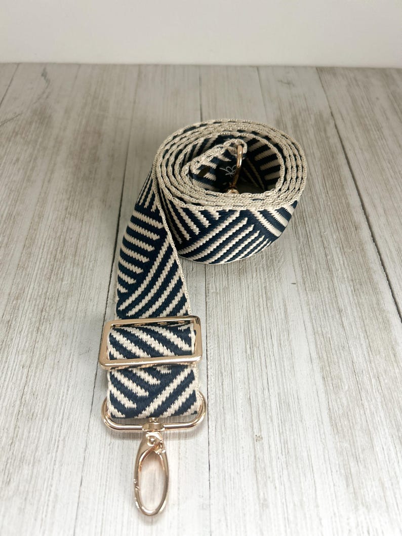 May include: A woven bag strap featuring a navy blue and cream chevron pattern. The strap includes a gold-toned metal adjustable slider and a swivel lobster clasp hook at the end, with the remaining length coiled into a circular shape.