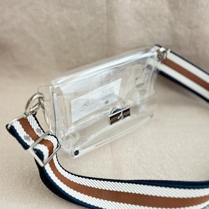 May include: A clear rectangular crossbody bag with a silver metal clasp closure. The bag features a wide adjustable strap with horizontal stripes in navy blue, white, and brown. A small white rectangular label is visible inside the transparent pouch.