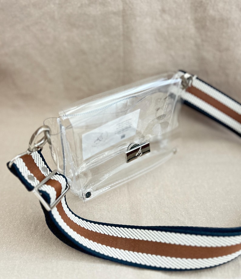 May include: A clear rectangular crossbody bag with a silver metal clasp closure. The bag features a wide adjustable strap with horizontal stripes in navy blue, white, and brown. A small white rectangular label is visible inside the transparent pouch.
