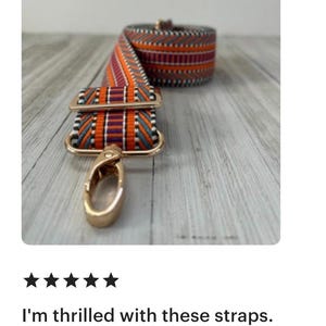 May include: A colorful woven bag strap with orange, purple, and teal geometric patterns. The strap has gold-colored metal hardware, including a swivel clasp and rectangular connectors. The strap is rolled up in the background.
