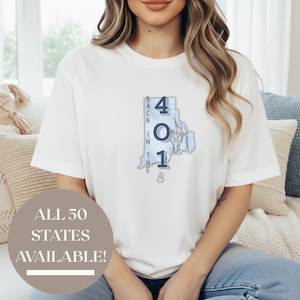 Puede incluir: Camiseta blanca con un gráfico del estado de Rhode Island y el texto "401" y "Back in the". La camiseta también tiene el texto "ALL 50 STATES AVAILABLE!" en un círculo.