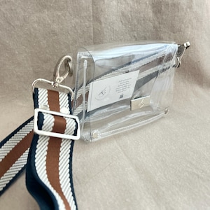 May include: A clear rectangular crossbody bag with silver-tone hardware and a detachable woven strap featuring navy blue, white, and brown stripes. A small white rectangular tag with black text and a logo is visible inside the transparent bag.